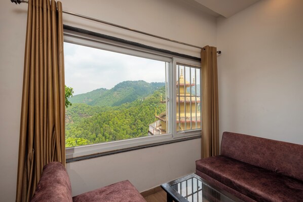 Una habitación con un gran ventanal que ofrece vistas a un área boscosa y un edificio con tejado dorado. Hay dos zonas de estar con cojines.