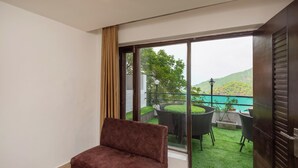 Ein modernes Zimmer mit einem braunen Sofa, großen Schiebeglasfenstern, die auf einen Balkon mit Sitzmöbeln und Blick auf eine grüne Landschaft führen.