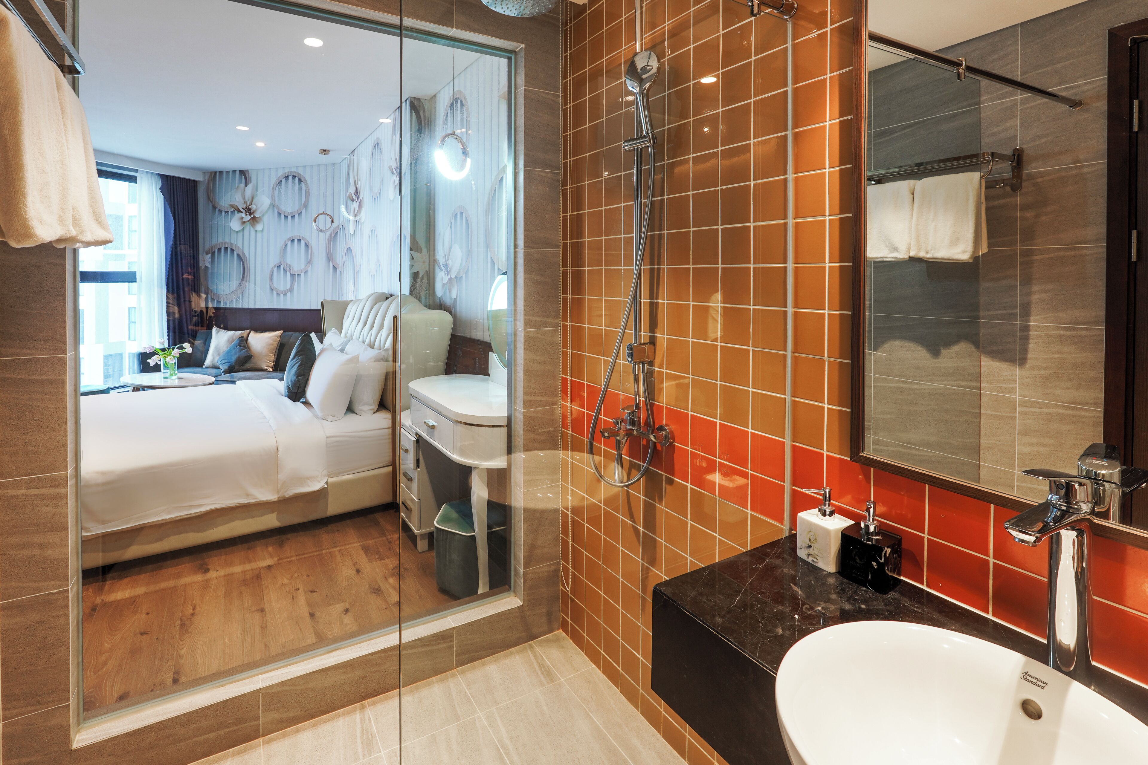 Quarto de hotel moderno com cabine de chuveiro de vidro, uma cama grande, banheiro com bancada preta e um espelho amplo.