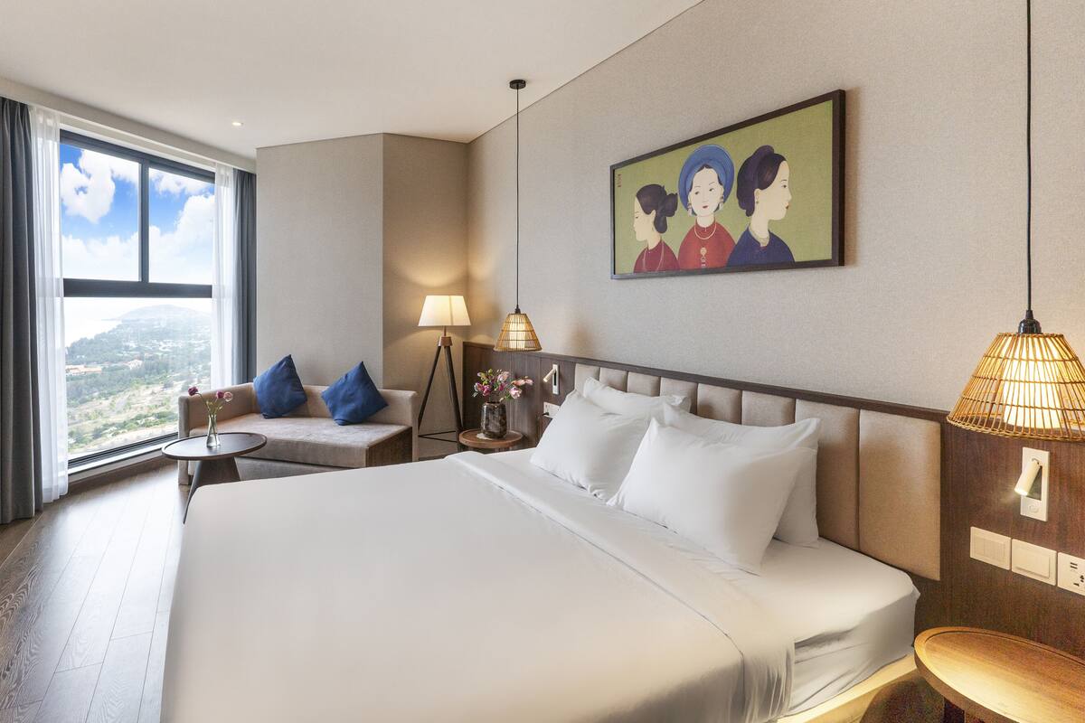 Une chambre d’hôtel avec un grand lit, un canapé, une petite table sur laquelle est posé un vase de fleurs, une lampe et un tableau au mur.