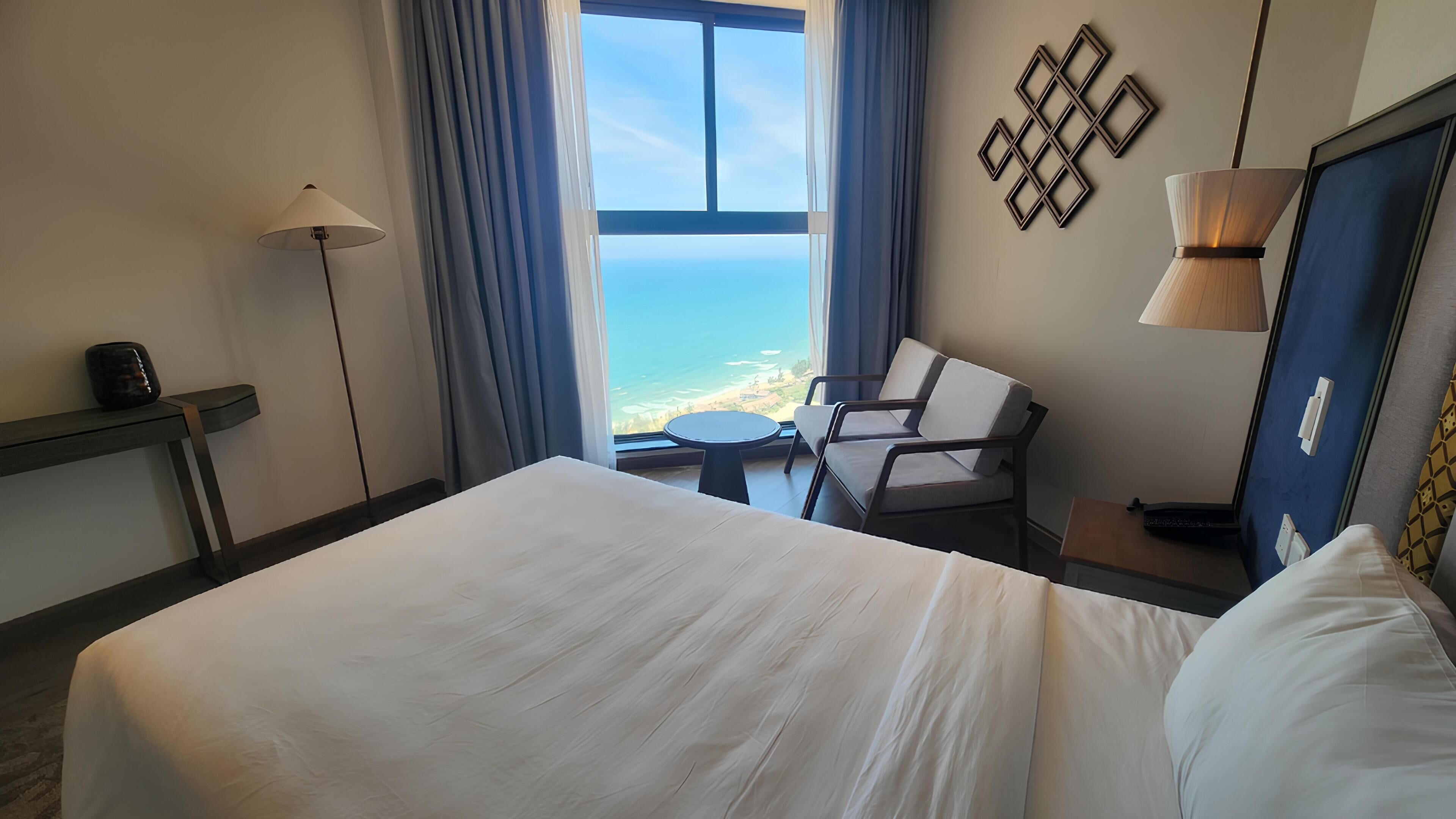 Habitación de hotel con una cama grande, una silla, una mesita, una lámpara y vistas al océano.