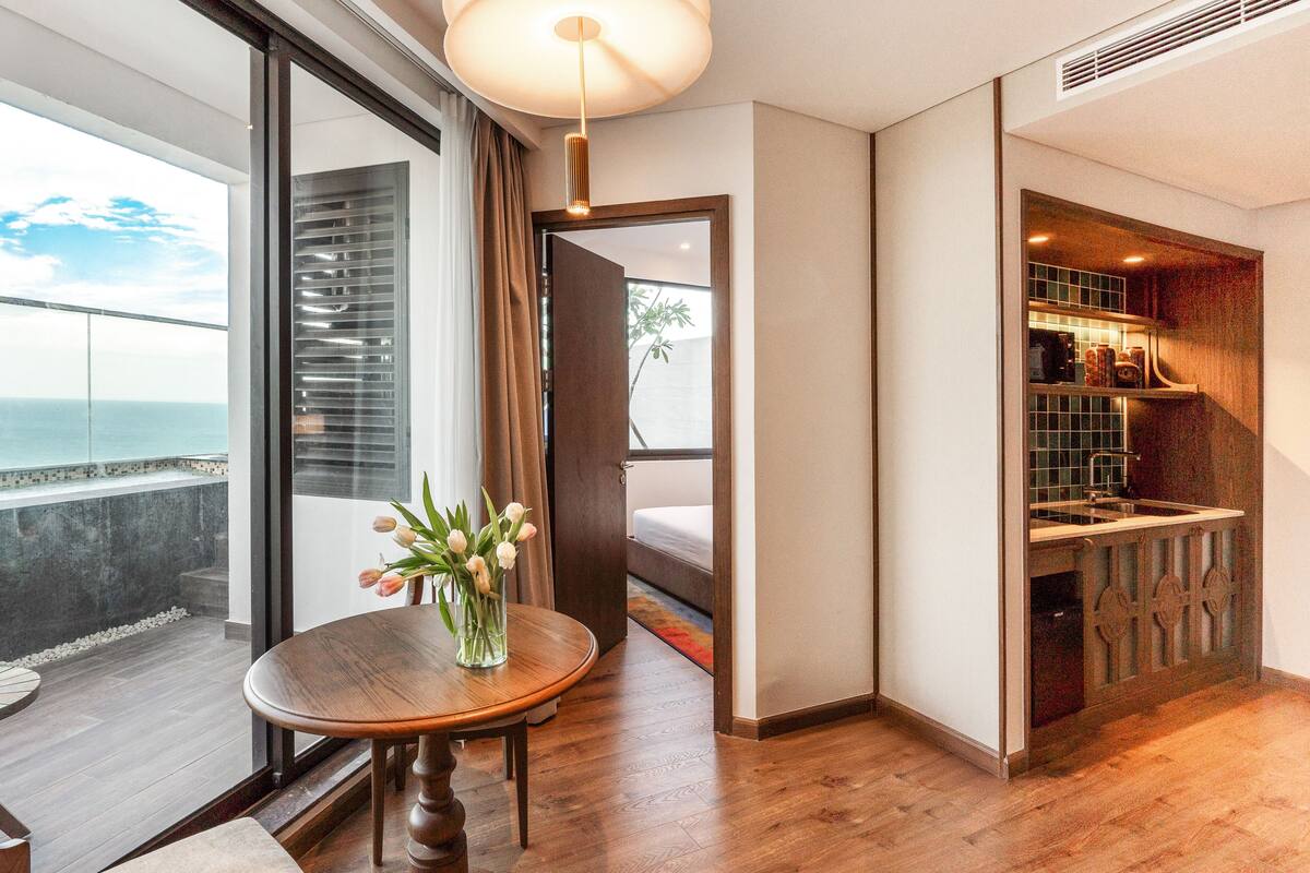 Une chambre d’hôtel moderne avec une grande fenêtre, une table à manger en bois ornée d’un vase de fleurs et un coin cuisine visible à travers une porte ouverte.