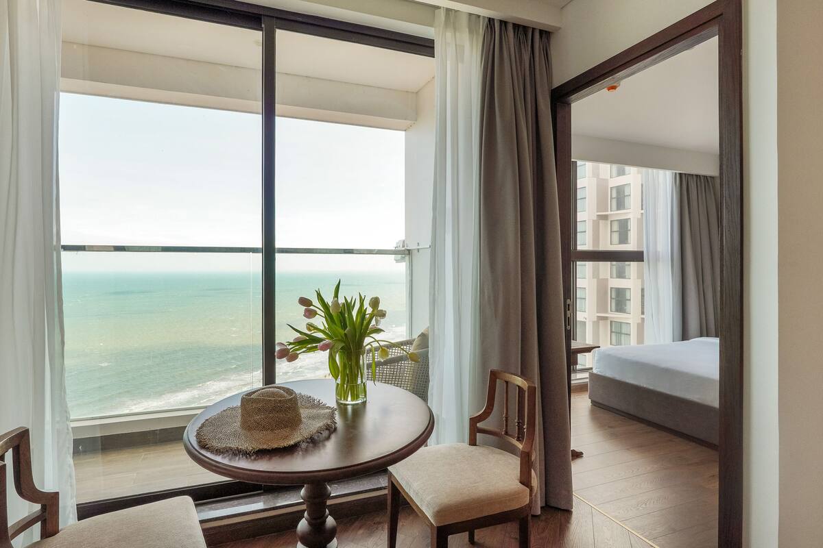 Une chambre d’hôtel avec une grande fenêtre, une table ronde surmontée d’un chapeau, une chaise, une petite plante et un lit.