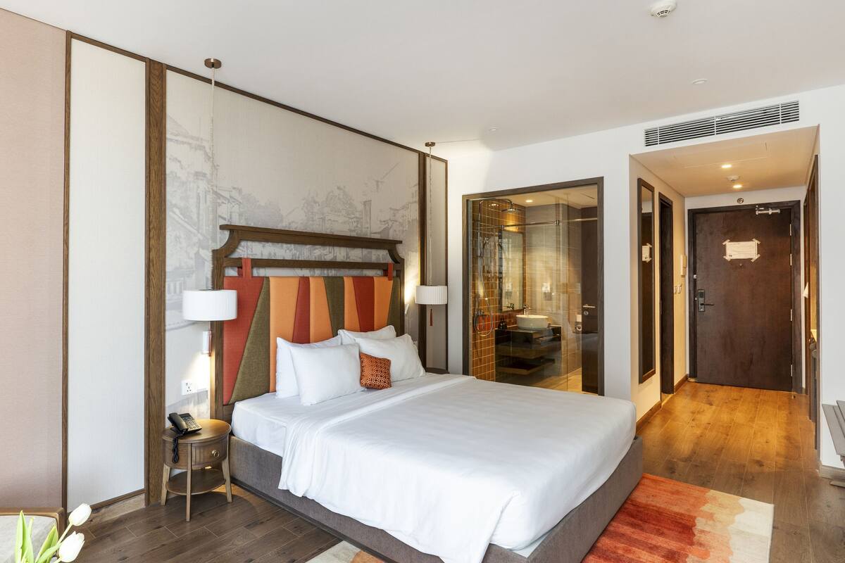 Une chambre d’hôtel moderne dotée d’un grand lit, d’une table de chevet avec une lampe, d’une douche en verre et d’une porte en bois avec une plaque numérotée.