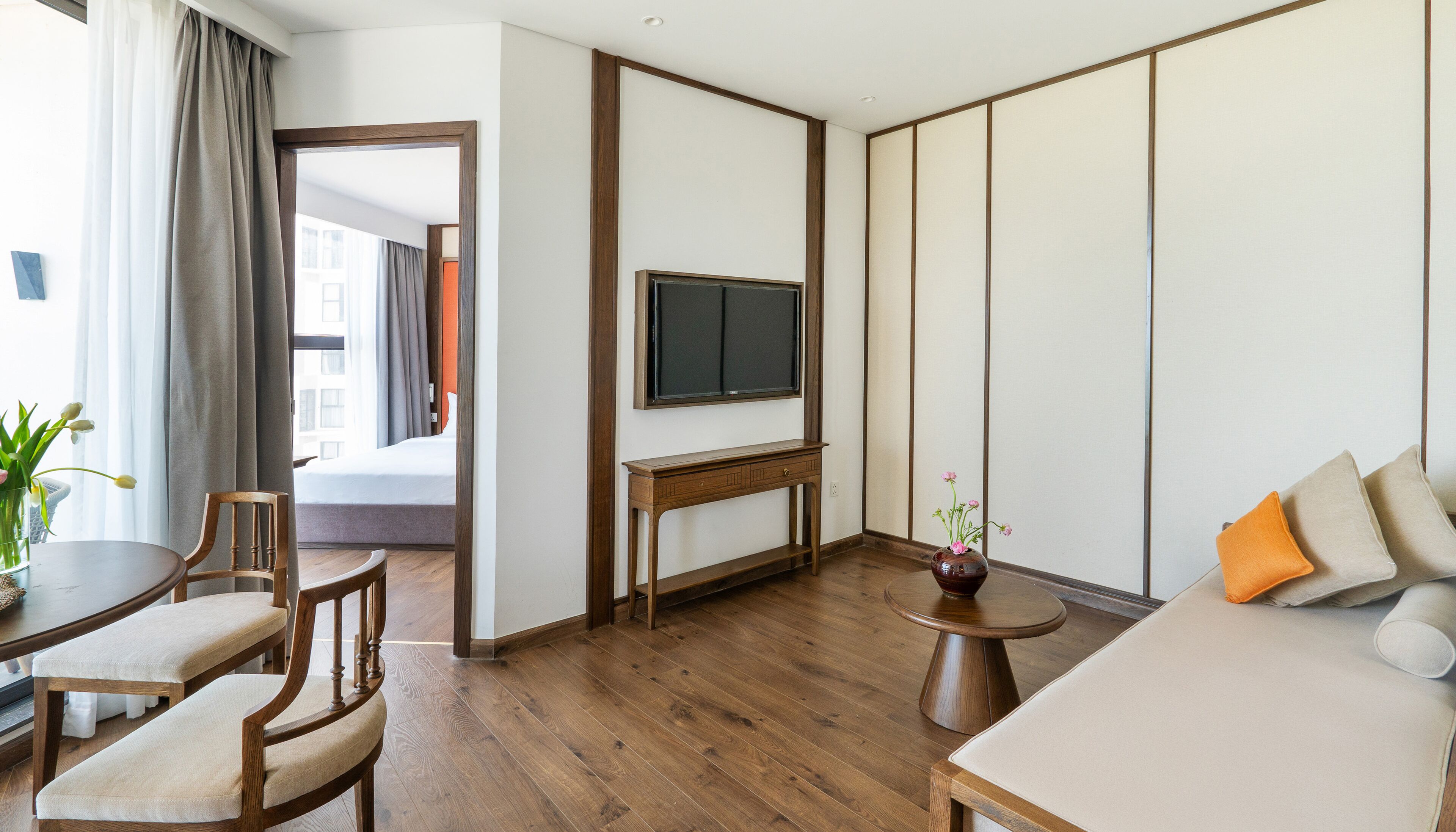 Habitación de hotel con piso de madera, cama, televisor sobre un soporte de madera, una mesita con un florero y un sofá con cojines.