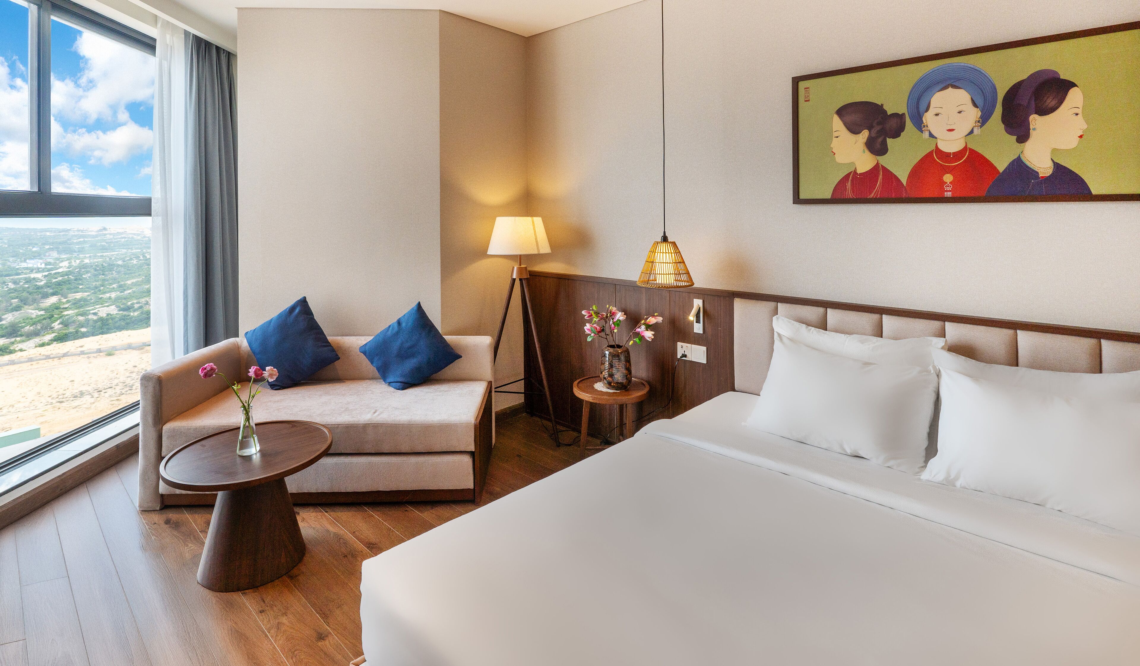 A hotel room with a bed, a sofa, a small table with a vase of flowers, a lamp, and a painting on the wall.