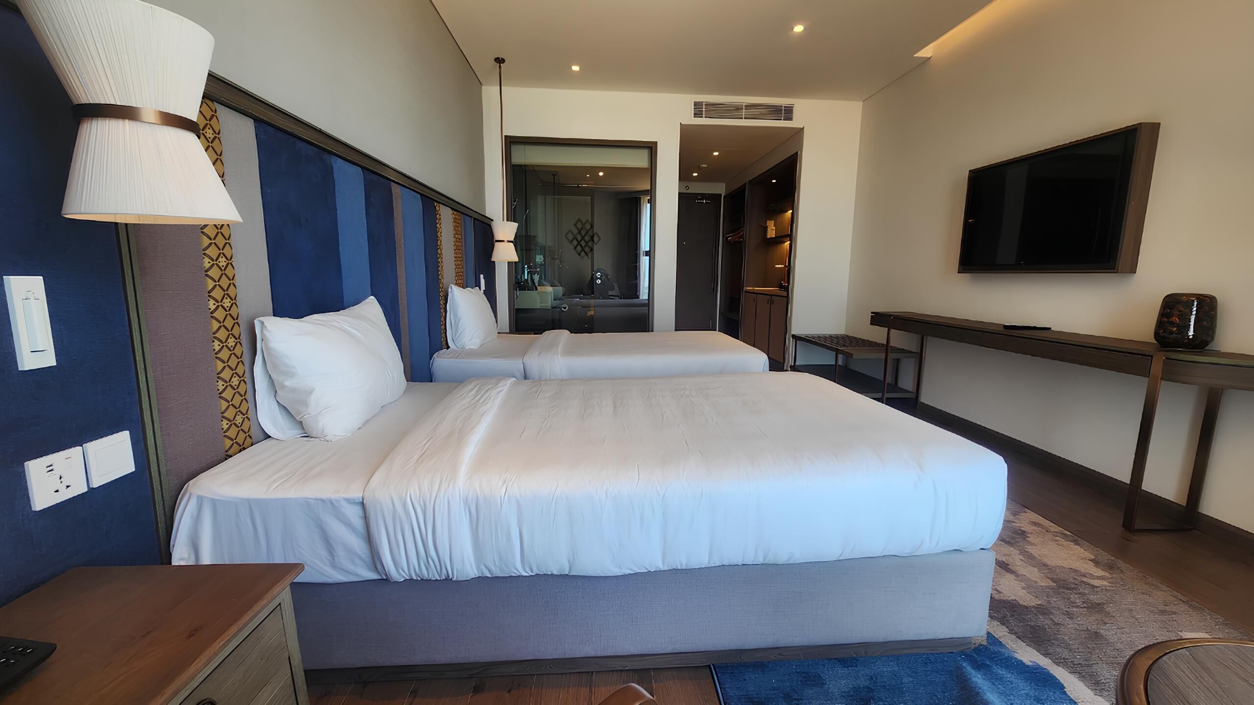 Habitación de hotel con dos camas, un escritorio, un televisor y un baño visible a través de una puerta abierta.