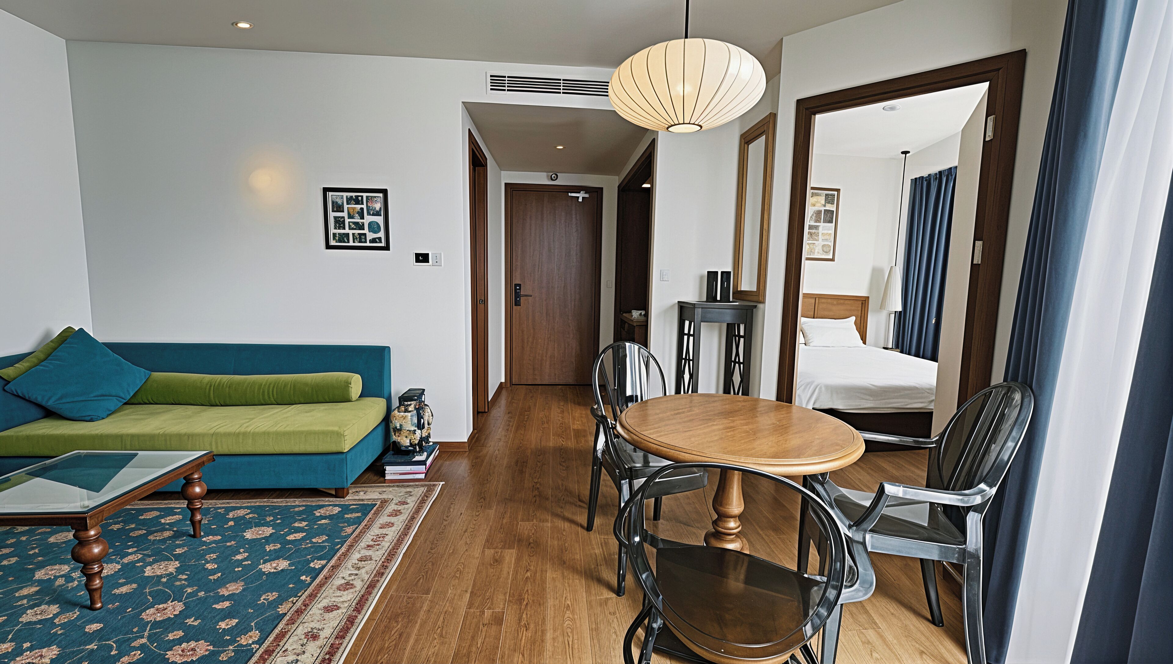 A hotel room with a green sofa, a wooden dining table with chairs, a bed with a wooden headboard, and a large window with curtains.