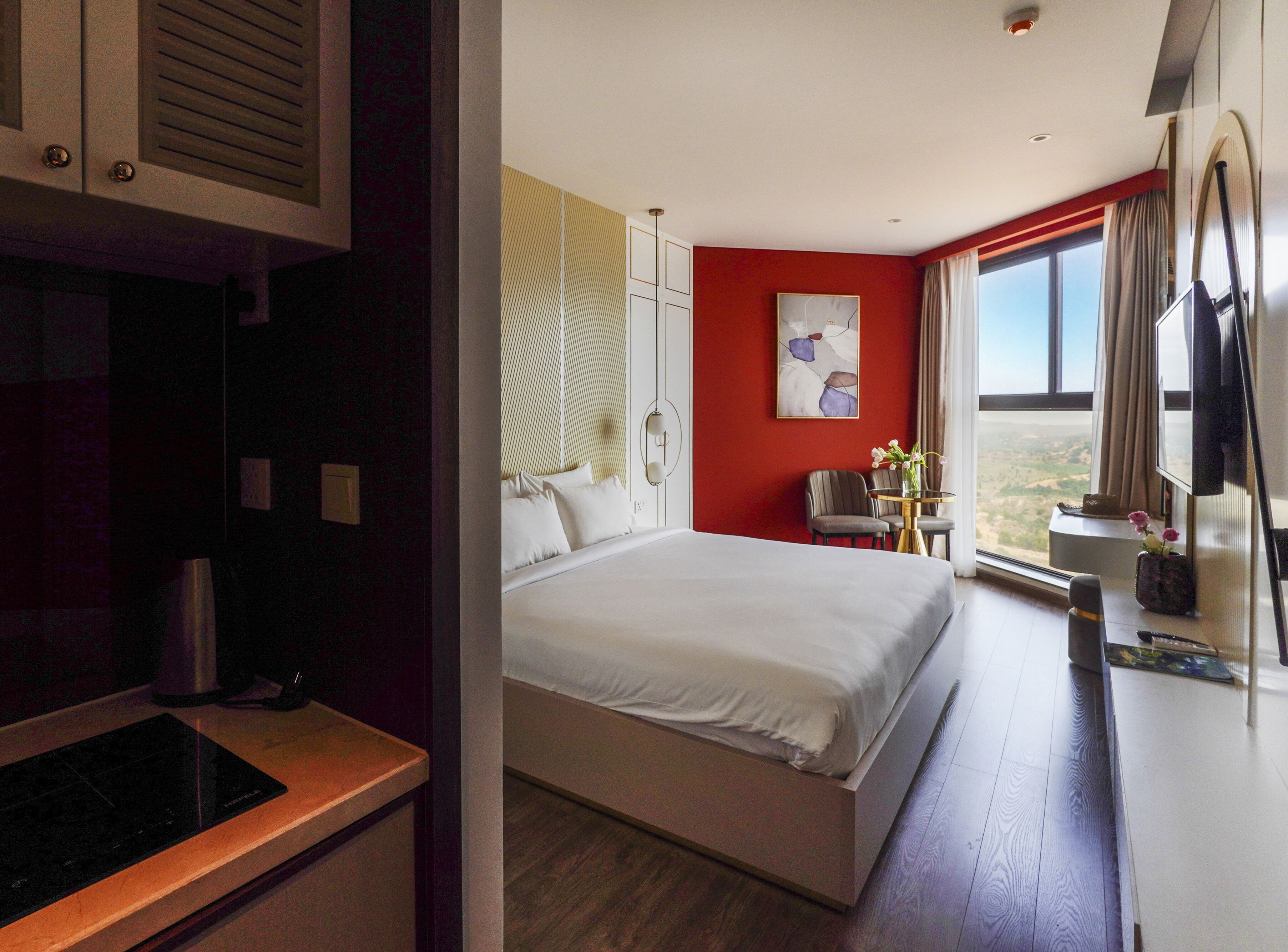 Quarto de hotel com cama, escrivaninha, cadeira, janela com vista e uma parede de destaque vermelha.