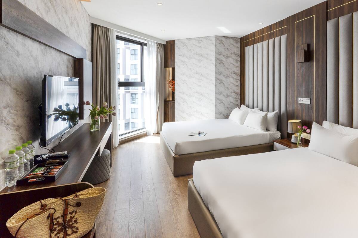 Une chambre d’hôtel moderne dotée d’un grand lit, d’un lit plus petit, d’un bureau avec une télévision et offrant une vue sur la ville grâce à de grandes fenêtres.