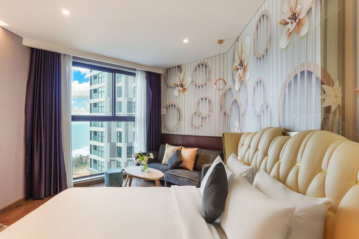 Une chambre d’hôtel avec un grand lit, un canapé, une petite table avec un vase, et une vue sur les bâtiments par la fenêtre.