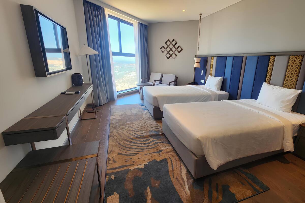 Une chambre d’hôtel avec deux lits, un bureau, une chaise, une grande fenêtre et un tapis à motifs.