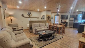 Una sala de estar acogedora con un conjunto de sofá, una mesa de centro y una zona de comedor con sillas. Se observa una cocina al fondo con un refrigerador y un fregadero.