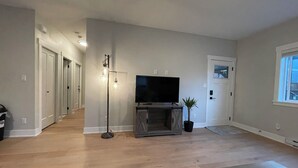 A living room with hardwood flooring, a flat-screen TV on a wooden stand, a floor lamp, and a potted plant. - Victoria luxury one bedroom suite (Langford)