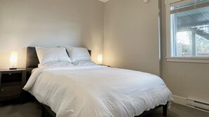A bedroom with a bed, two bedside tables, and a window with a view of greenery. - Victoria luxury one bedroom suite (Langford)
