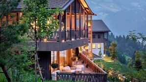 A modern two-story house with a balcony, surrounded by lush greenery and mountains in the background. - AMONTI Chalets (Campo Tures)