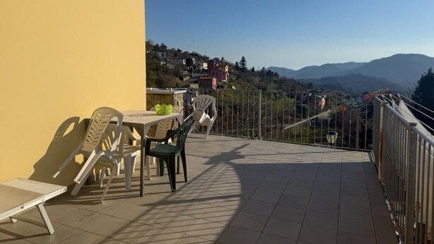 A balcony with a table and chairs, a view of a hillside with buildings, and a clear sky. - IL FILO DI ARIANNA (Ricco del Golfo di Spezia)