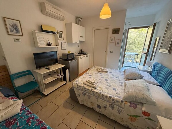Een compacte slaapkamer met een bed, een bureau met computer, een klein keukentje en een balkon.