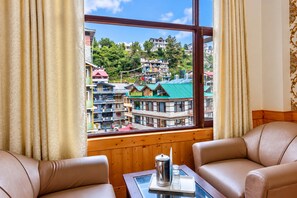 A room with two leather sofas, a wooden table with a coffee pot and glasses, and a window with a view of a town with colorful buildings. - Hotel Day Night (Manali)