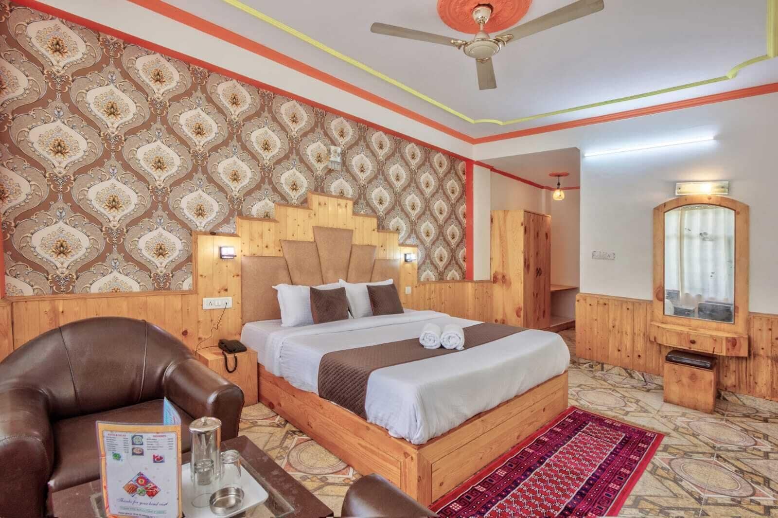 A hotel room with a large bed, wooden headboard, patterned wallpaper, a ceiling fan, a mirror, and a red carpet.