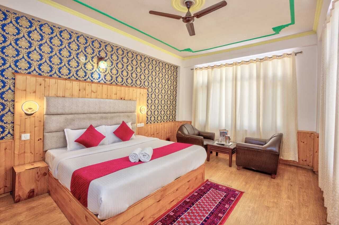 A hotel room with a wooden headboard, a large bed with red pillows, a small table, two chairs, and a ceiling fan.