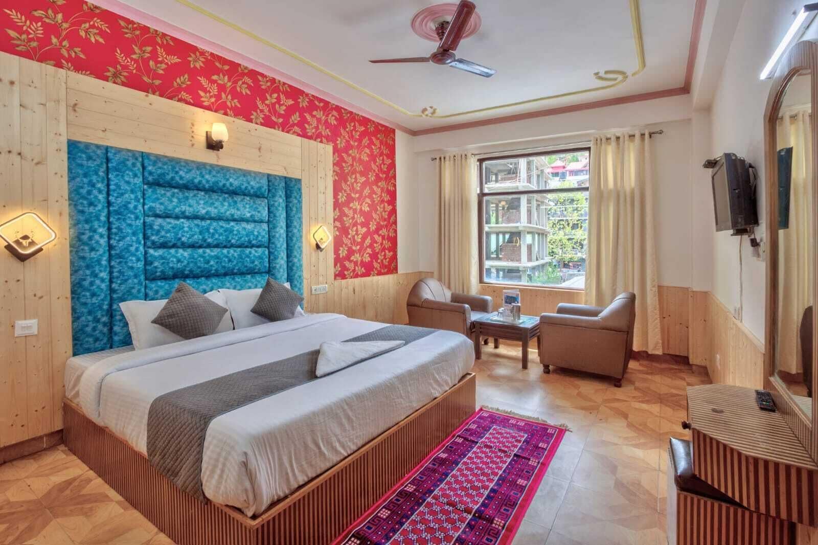 Habitación de hotel con una cama grande, cabecera de madera, pared con estampado floral rojo, ventilador de techo, ventana con cortinas y una mesita con una silla.