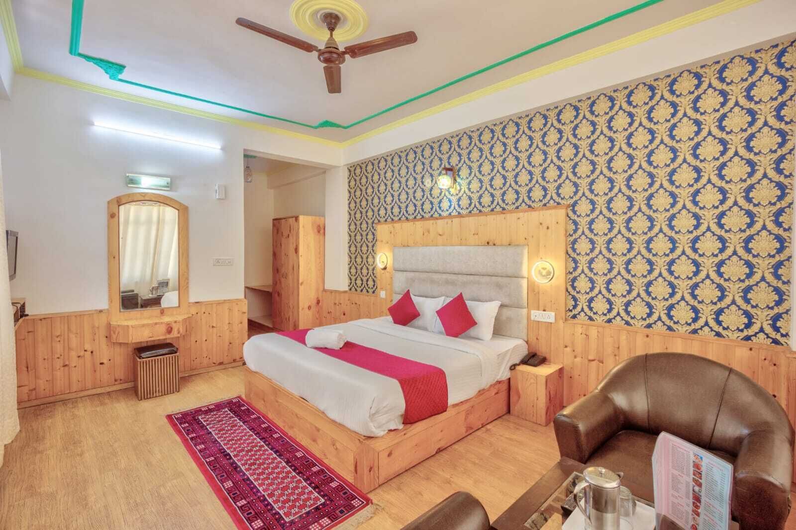 Une chambre d’hôtel avec une tête de lit en bois, un lit avec une literie blanche et rouge, un plancher en bois, un tapis à motifs rouges, un fauteuil en cuir marron et un ventilateur de plafond.