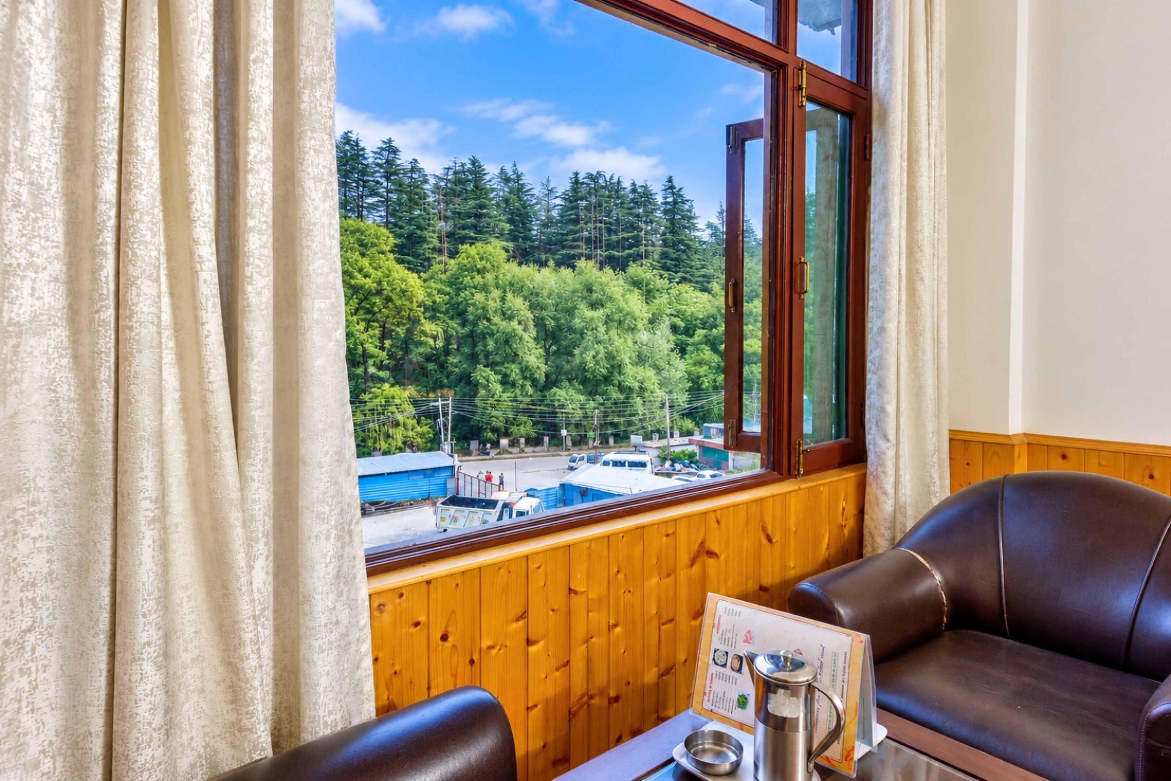 A room with a view of a forest and a road, a wooden wall, a leather sofa, and a table with a menu and a coffee pot.