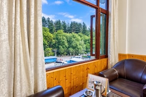 A room with a view of a forest and a road, a wooden wall, a leather sofa, and a table with a menu and a coffee pot. - Hotel Day Night (Manali)