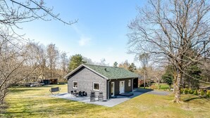 A small, single-story cabin with a green roof, a patio area with outdoor seating, and a well-maintained lawn.
