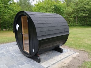 A small, black, barrel-shaped structure with a wooden door and a window, situated on a paved area with grass and trees in the background.