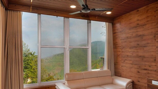 A room with a large window, a sofa, a table, and a ceiling fan. - Mountain Villa Mussoorie  (Mussoorie)