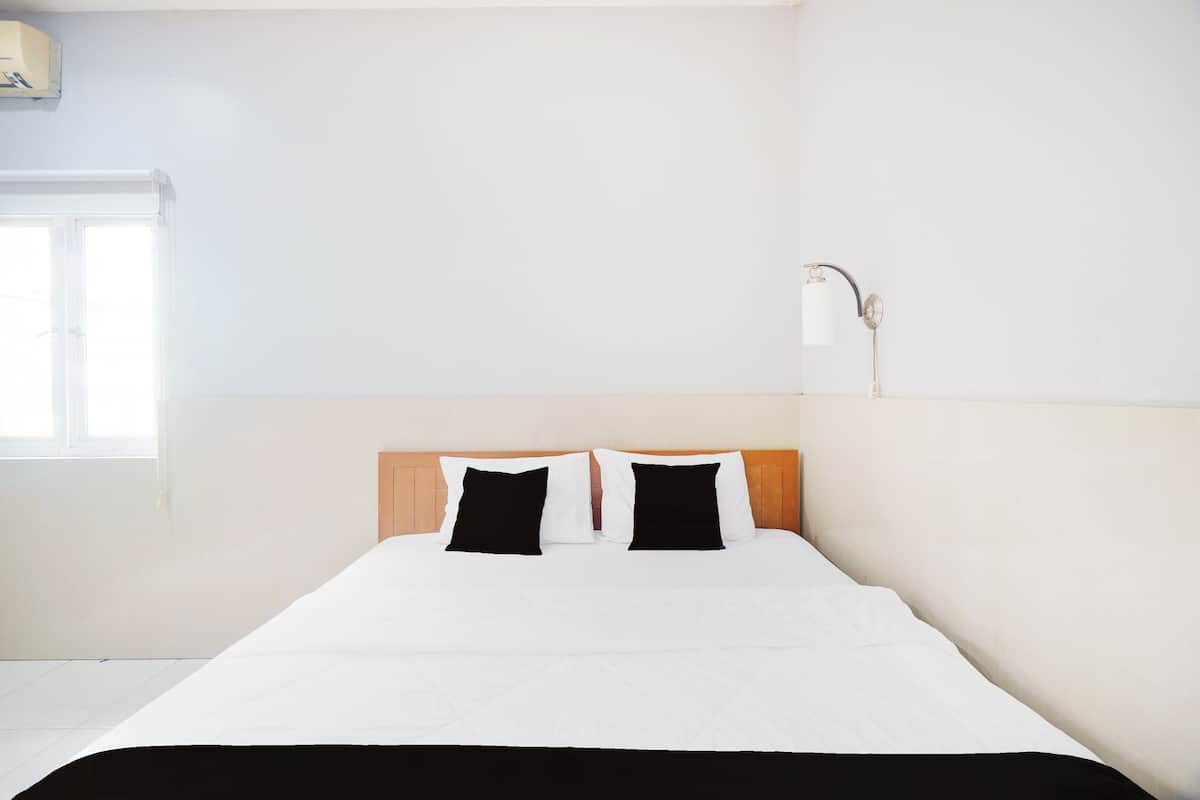 Une chambre d’hôtel avec un lit, deux oreillers noirs et blancs, une tête de lit en bois, un mur blanc et une fenêtre.