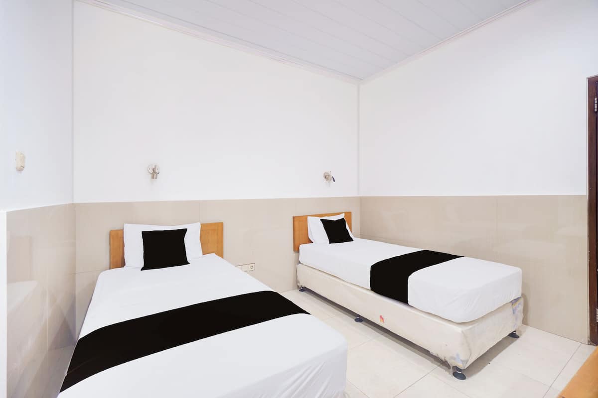 Une chambre d’hôtel avec deux lits simples, chacun doté d’une tête de lit en bois, de draps blancs et de coussins noirs décoratifs. La chambre a un sol carrelé et des murs blancs.