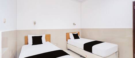 Habitación de hotel con dos camas individuales, cada una con cabecero de madera, ropa de cama blanca y almohadas negras decorativas. El piso es de baldosas y las paredes son blancas.