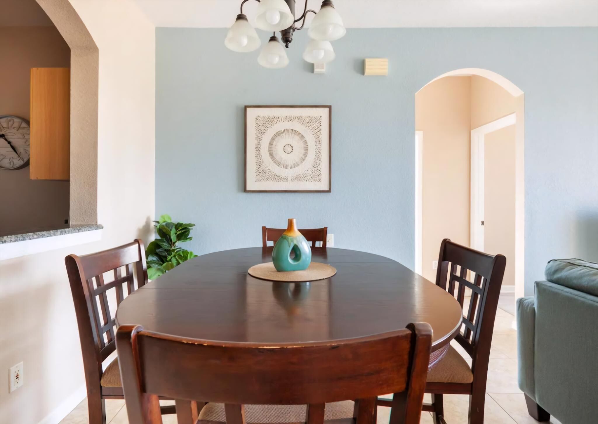 A dining area with a round wooden table, six chairs, a teal vase, and a framed artwork on the wall.