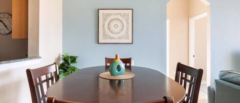 A dining area with a round wooden table, six chairs, a teal vase, and a framed artwork on the wall.