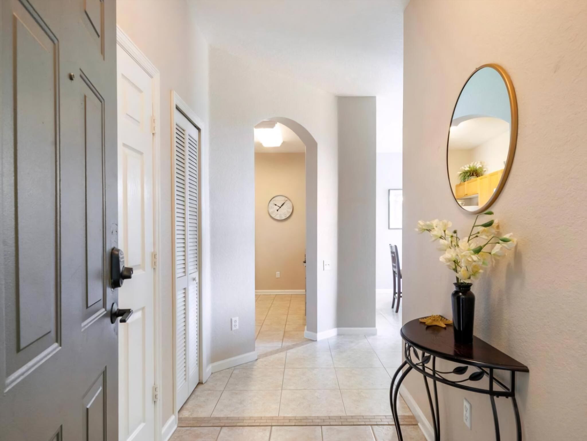 A hallway with a door, a wall-mounted clock, a mirror, and a small table with a vase of flowers.