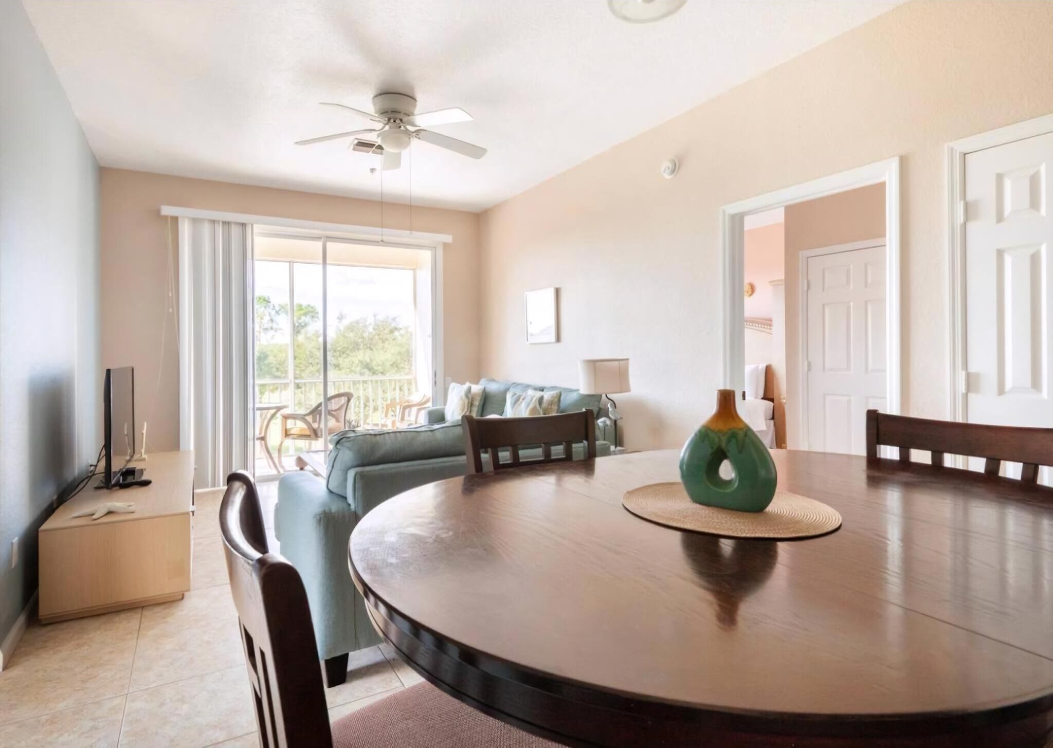 A dining area with a round table, chairs, a ceiling fan, and a view of the outdoors through large windows.