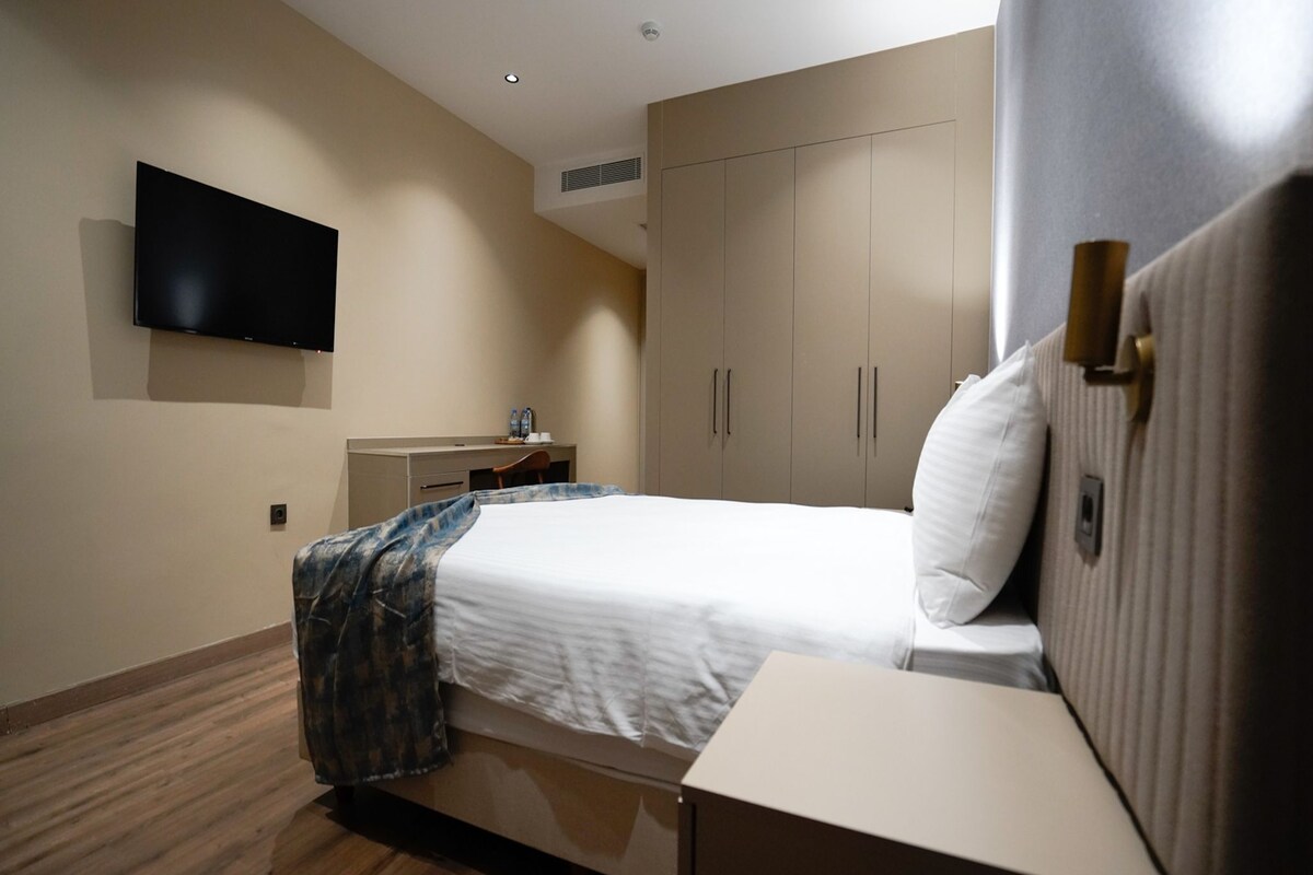A hotel room with a bed, a desk, a flat-screen TV mounted on the wall, and a wardrobe.