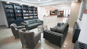 A modern lobby with a grey sofa, patterned armchairs, a coffee table, and a bookshelf filled with books and awards. - Zade Life OTEL (Gaziantep)