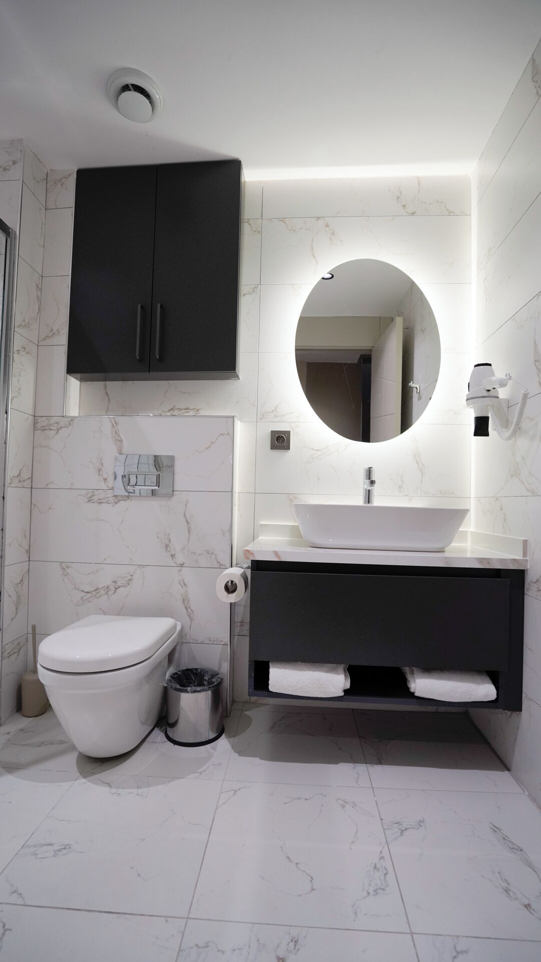 A modern bathroom with a white toilet, black cabinets, a round mirror, and a white sink.
