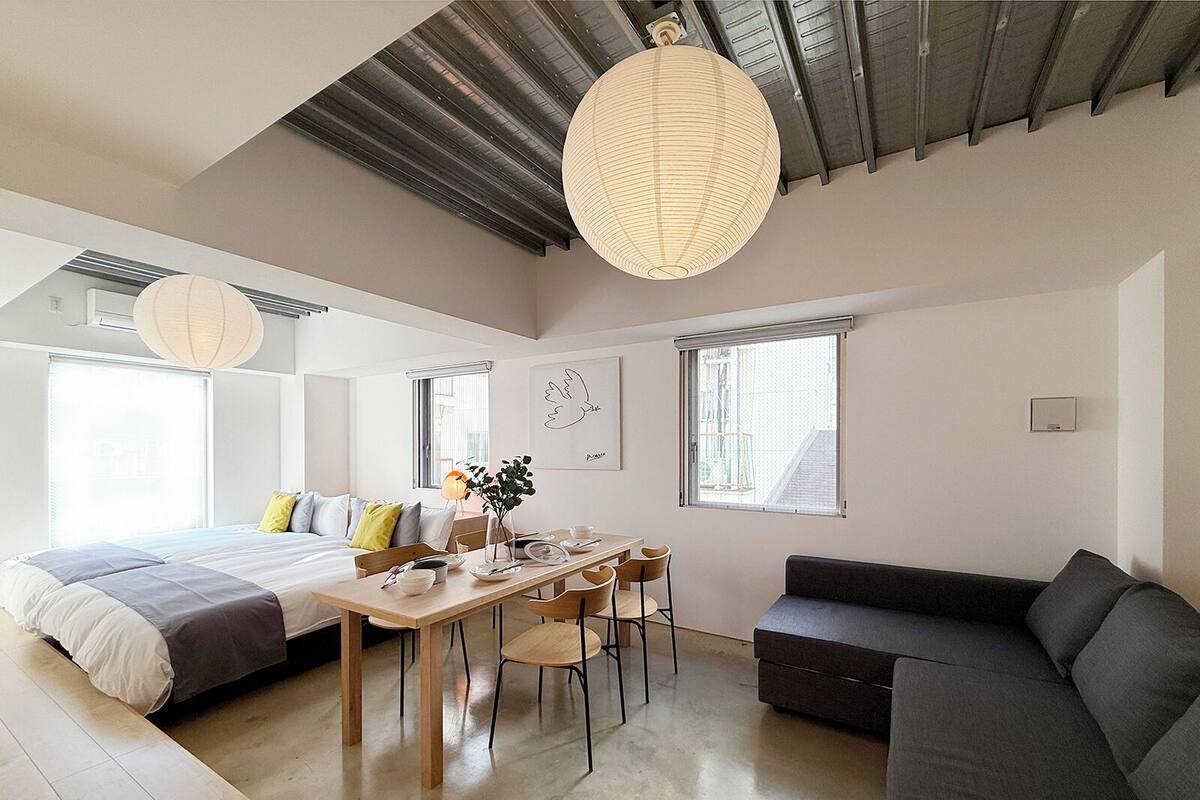 A modern, well-lit room with a bed, a sofa, a dining table, and chairs. There are windows providing natural light and a view of the outside.