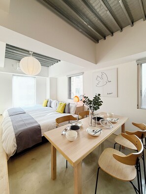 A modern, well-lit room with a large bed, a dining table with chairs, and a minimalist decor including a wall art piece and a hanging light fixture. - Arts Dobashi (Hiroshima)