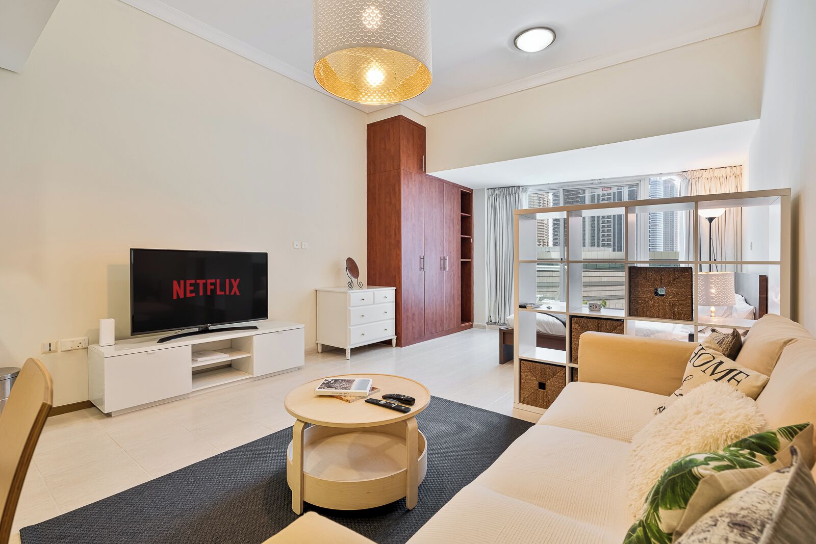 Ein modernes Wohnzimmer mit einem Flachbildfernseher, der 'NETFLIX' anzeigt, einer weißen Unterhaltungsanlage, einem Holztisch, einer Couch mit 'HOME'-Kissen und einem großen Fenster mit Blick auf die Stadt.