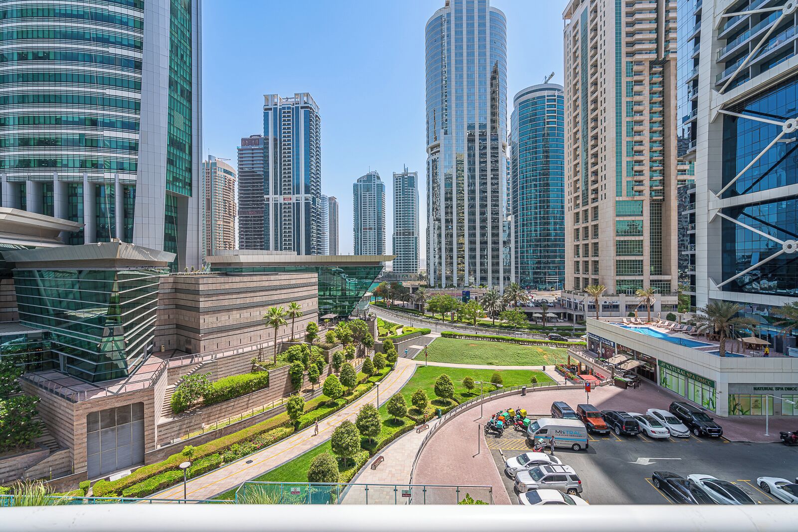 A modern urban area with high-rise buildings, a central green space, and a parking area with vehicles.