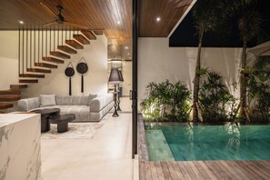 Ein modernes Interieur mit einer Holztreppe, einem grauen Sofa und einem Poolbereich mit tropischen Pflanzen.