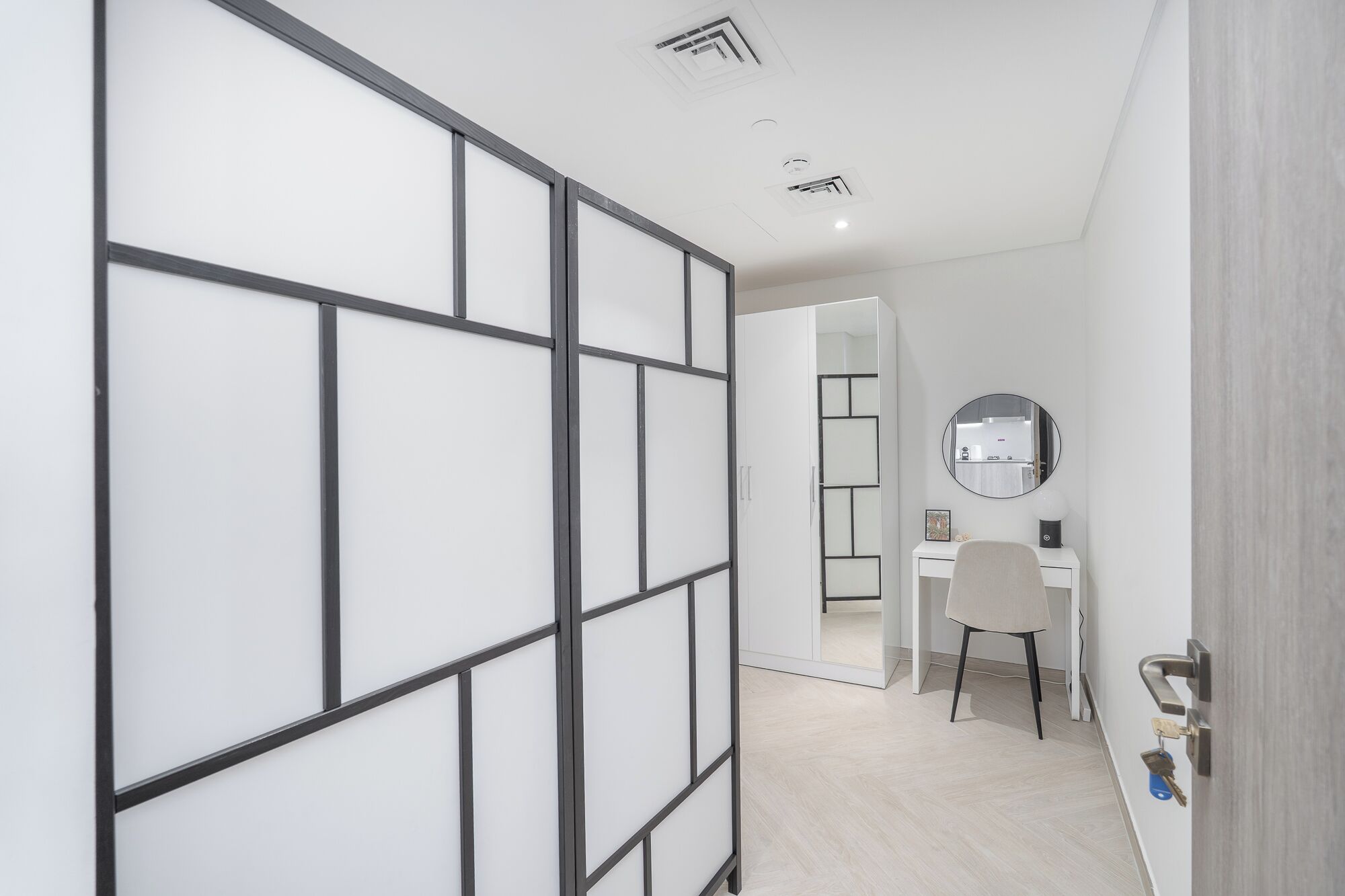 A modern, minimalist hallway with a white wardrobe, a desk with a chair, and a round mirror on the wall.
