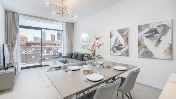 A modern dining area with a long table set for four, a sofa, and a city view through large windows.