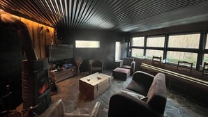 Un intérieur chaleureux avec une cheminée en pierre, des sièges confortables et de grandes fenêtres offrant une vue sur l’extérieur.